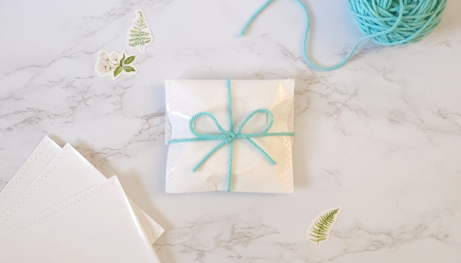 Styled gift wrap scene on a white marble surface. In the middle is a small gift wrapped in white paper with a turquoise bow. Also shown are white gift bags, a blue skein of yarn and botanical stickers.