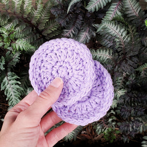 A hand holds a set of 2 purple crochet car coasters in front of green ferns.
