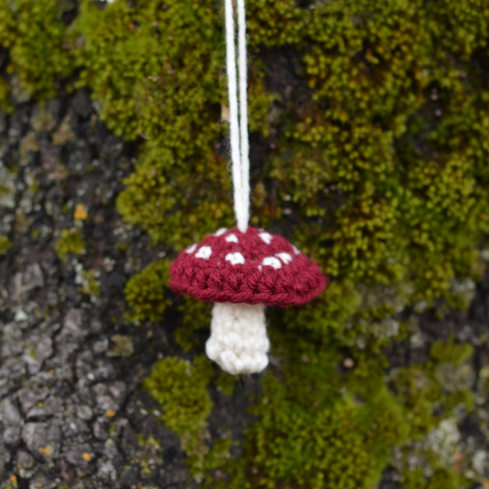 Small crocheted red and cream mushroom hanging in front of a moss covered tree.